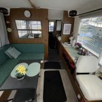 Photo de l'intérieur du bateau depuis la porte d'entrée. On voit à gauche une banquette avec une table rectangulaire grise, une fleur jaune et deux