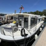 Vue de côté du bateau avec sa coque blanche et noire. On voit le pont avant avec une table