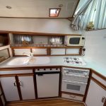Photo de la cuisine du bateau. Elle est blanche avec les contours des meubles en bois. On voit un évier blanc à gauche, une plaque de cuisson au gaz, un four, un réfrigérateur et un micro-ondes à droite.
